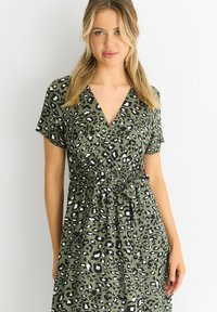 Green leopard print dress with short sleeves, a deep V-neck, cinched waist, and a tie detail at the waist. Soft fabric texture.