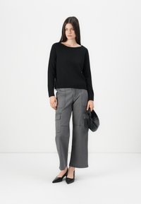 Femme portant un top noir à manches longues, un pantalon cargo large gris, des escarpins noirs à bride arrière, tenant un petit sac à main noir, se tenant sur un fond blanc.