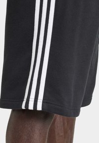 Černé sportovní šortky z měkké látky s třemi svislými bílými pruhy po straně. Lem je prošitý pro větší odolnost.