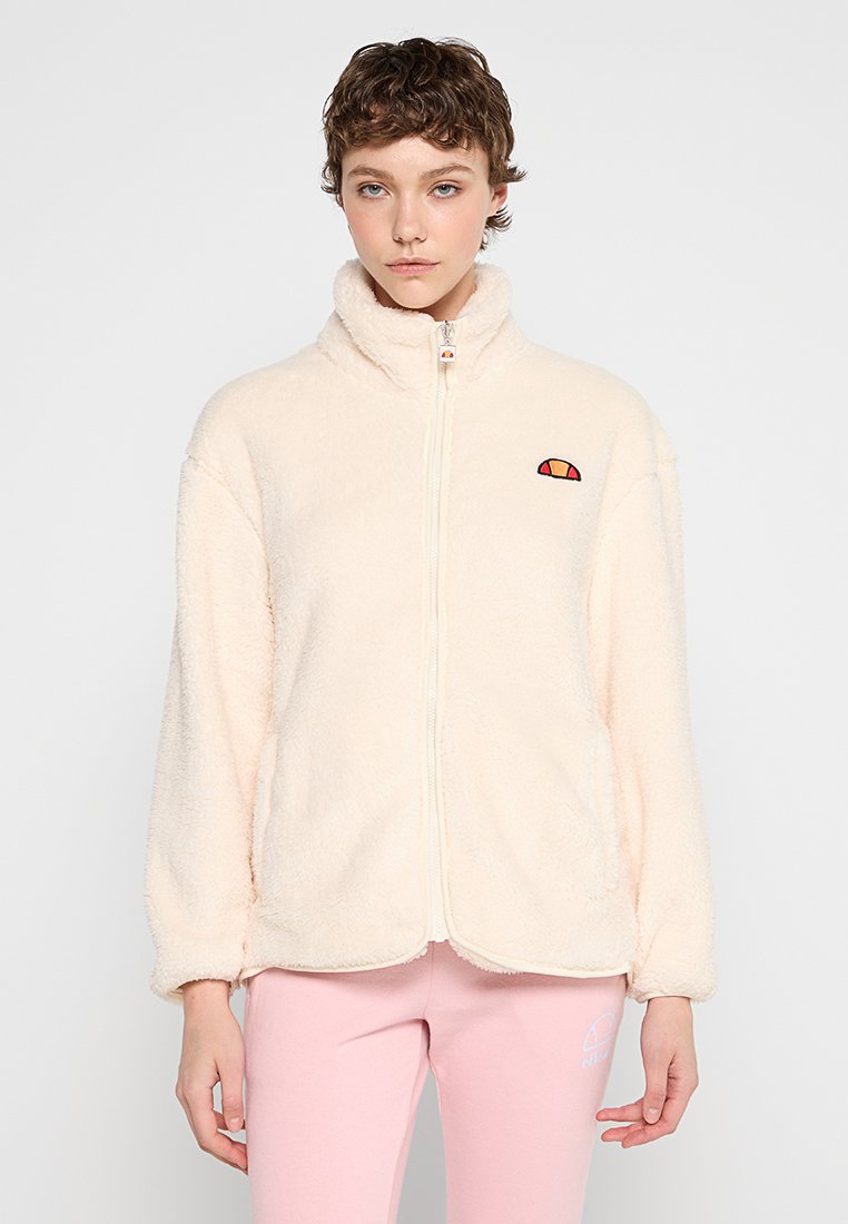 Veste en polaire de couleur crème douce, dotée d'un col montant, d'une fermeture éclair à l'avant et d'un petit logo brodé en orange et rouge sur la poitrine.