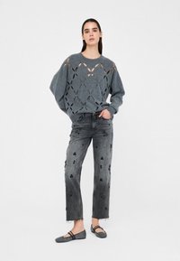 Maglione grigio a maglia con dettagli ritagliati, abbinato a jeans grigi a vita alta con motivi decorati. Include anche scarpe flat.