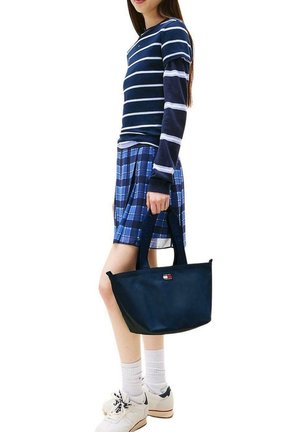 Mujer con blusa de rayas azules y mangas largas, falda a cuadros azul, que lleva un bolso tote azul marino, calcetines blancos y zapatillas blancas.