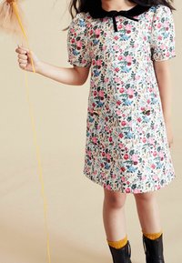 Robe fleurie avec des manches bouffantes courtes, fond blanc, fleurs roses et bleues, col en velours noir, deux poches, associée à des bottes noires et des chaussettes jaunes.