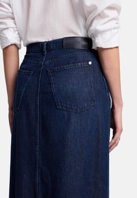 Gonna in denim scuro con vita alta, dotata di due tasche posteriori, un'etichetta in vita e una superficie liscia. Il tessuto presenta un aspetto denim classico.