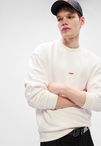 Λευκό φούτερ με ριγέ μανίκια και τελείωμα, διακοσμημένο με κόκκινο λογότυπο "HUGO" στο στήθος. Λείο ύφασμα και χαλαρή εφαρμογή. Μαύρη ζώνη ορατή από κάτω.