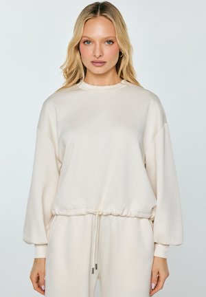 Blonde sieviete, tērpusies brīvā, garām piedurknēm bēša krāsas sporta krekla un atbilstošu bēša krāsas biksēs ar aukliņu viduklī, stāv pret vienkrāsainu fonu.