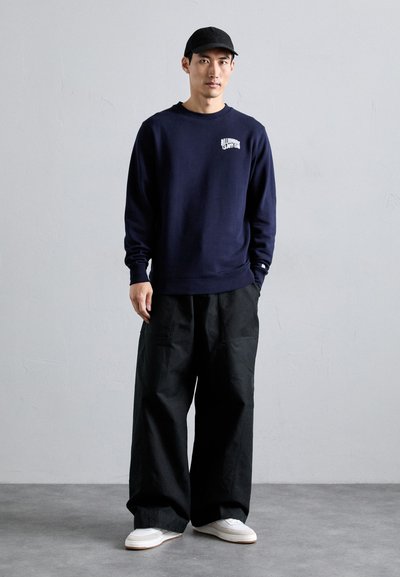 Marineblå genser med frontlogo, svarte vide bukser og hvite sneakers. Modellen har på seg en svart caps. Enfargede plagg med minimale detaljer.