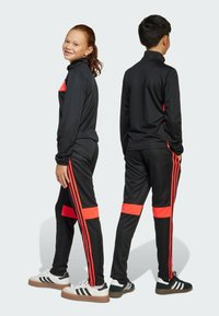 Svart träningsdräkt med en zip-up jacka med röda detaljer och matchande byxor, båda med röda ränder på sidorna. Vita sneakers med svarta detaljer.