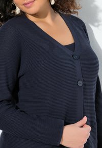 Cardigan bleu marine en tissu côtelé texturé, avec un décolleté en V et deux boutons à l'avant, manches longues et finition douce.