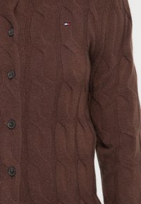 Brun strikket cardigan med kabelemønster, fem svarte knapper og en liten rød, hvit og blå logo på brystet.