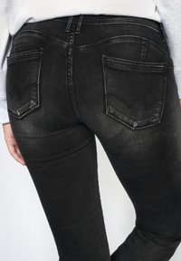 Jeans en denim noir avec un design ajusté, style à cinq poches et des accents de couture visibles sur les poches arrière.