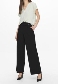 Lichtgroene v-hals top en zwarte wijde broek met een knop, weergegeven op een model. De broek heeft zijzakken en een gladde stof.