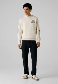 Sweatshirt creme com um logótipo no peito, apresenta texto colorido na manga direita. Combinado com jeans escuros e ténis brancos.