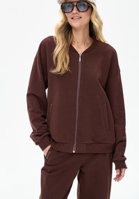 Brown zip-up sweatshirt with dropped shoulders, ribbed cuffs, and front pockets. Worn with matching pants, features brown fabric and subtle branding.