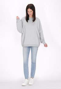 Szürke, oversized kapucnis pulcsi hosszú ujjakkal és bordás mandzsettákkal, világoskék skinny farmerral és fehér sportcipővel egy semleges háttér előtt.