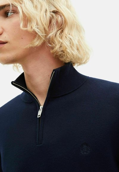 Granatowy wełniany sweter z wysokim kołnierzem i srebrnym zamkiem. Posiada subtelną detalizację logo na piersi. Miękka tekstura.