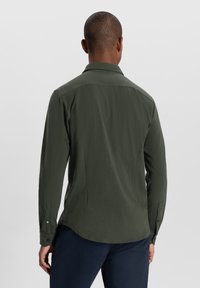 Cămașă cu mâneci lungi de culoare verde închis, cu guler, având o textură fină și manșete cu butoni. Spatele prezintă o tăietură dreaptă și cusături subtile.