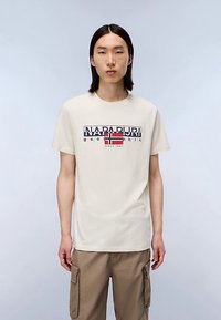Beige katoenen T-shirt met een marineblauw en rood grafisch ontwerp met de tekst "NAPAPIJRI geographic". Korte mouwen en ronde hals, ontspannen pasvorm.