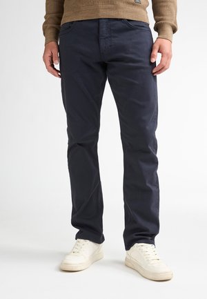 Hombre con pantalones azul marino, suéter marrón y zapatillas blancas, de pie frente a un fondo sencillo.