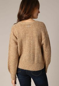 Beige gebreide trui met een gestructureerd diamanten patroon, wijde mouwen en een geribbelde zoom, met een kleine decoratieve knop op de achterkant.