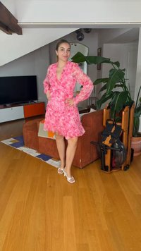 Γυναίκα με ροζ φλοράλ φόρεμα και λευκά τακούνια στέκεται με τα χέρια στη μέση σε ένα καθιστικό με τηλεόραση, μεγάλο φυτό και μηχάνημα κωπηλασίας.