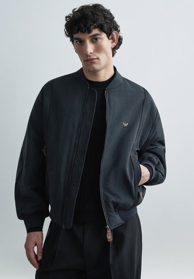 Blouson aviateur noir avec une finition texturée, des poignets côtelés et une fermeture éclair. Comprend un petit emblème sur la poitrine et des poches latérales.