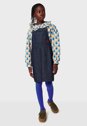 Robe salopette en denim avec un motif à rayures, chemise à manches longues à pois bleus et jaunes, leggings violets avec des accents verts et chaussures brunes.