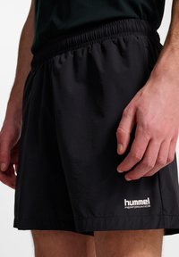 Schwarze sportliche Shorts aus leichtem Stoff mit einem elastischen Bund und einem weißen Logo am Saum. Glatte Textur, knielange Passform.