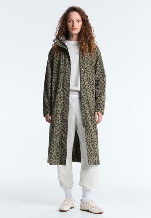 Junge Frau steht und trägt einen langen, grünen Mantel mit Leopardenmuster über einem hellgrauen Sweatshirt und einer Jogginghose sowie weißen Turnschuhen.