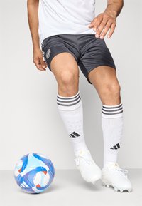 Jucător de fotbal în ghete albe, pantaloni scurți negri și șosete albe, controlând o minge de fotbal cu un model albastru și alb pe un fundal simplu.