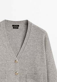 Vaaleanharmaa V-puolikkaa villa- ja kashmir-neulecardigan, jossa on kaksi metallinappia ja ribbaustekniikalla tehty taskut edessä.