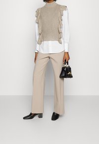 Beige neuleliivi, jossa röyhelökoristeita valkoisen napitettavan paidan päällä. Kermanväriset leveälahkeiset housut ja mustat korolliset mules-kengät. Pieni musta käsilaukku.