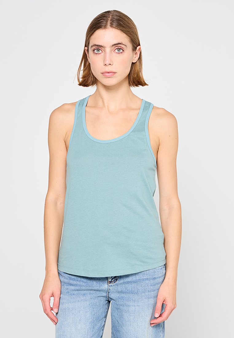 Jeune femme avec des cheveux châtain clair portant un haut sans manches bleu clair et un jean bleu devant un fond blanc uni.