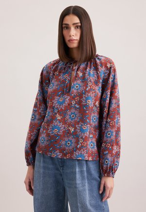 Langärmlige Bluse mit rundem Ausschnitt, die ein florales Muster in Blau und Weiß auf einem rostfarbenen Hintergrund aufweist. Weicher Stoff mit elastischen Manschetten.