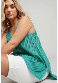 Tyrkis stickad ärmlös topp med ett vågigt mönster, med en lös passform och sidslitsar, i kombination med vita shorts.