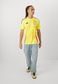 Yellow sports jersey with purple and orange accents, featuring a team logo and short sleeves. Worn with light blue jeans and sneakers.