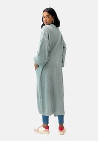 Lång, ljus mintgrön stickad cardigan med vida ärmar och sidofickor, bärs över åtsittande jeans och vita sneakers med röda detaljer.