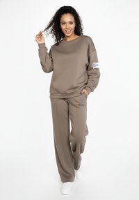 Brązowa bluza i dopasowane, szerokie spodnie wykonane z gładkiej tkaniny. Bluza ma metkę na rękawie. Dopełnieniem stroju są sneakersy.