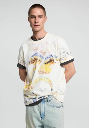 Junger Mann mit kurzen Haaren, der ein cremefarbenes T-Shirt mit bunten abstrakten Mustern über einem schwarzen Langarmshirt und hellen Jeans trägt.