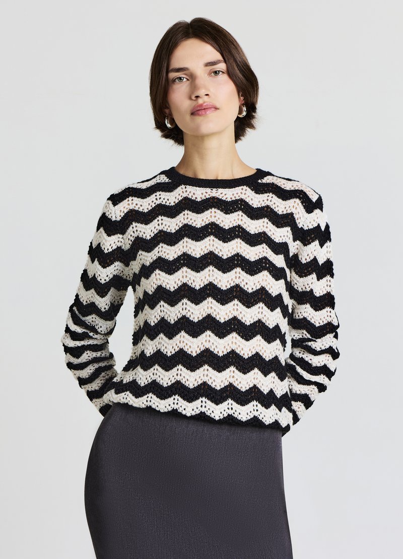 Pull en tricot avec un motif en zigzag noir et blanc, présentant un col rond et des manches longues, porté par-dessus un bas foncé et ajusté.