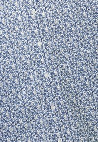 Camicia bianca decorata con un motivo floreale blu, caratterizzata da piccoli fiori blu, con bottoni bianchi lungo la parte anteriore per la chiusura.