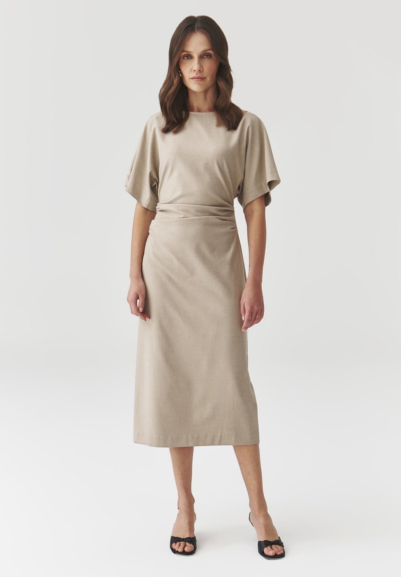 Beige midi mekko, jossa on lyhyet, leveät hihat ja vyötärö kireällä. Sileä tekstuuri, yksinkertainen muotoilu ilman kuvioita. Yhdistetty mustiin korkokenkiin.