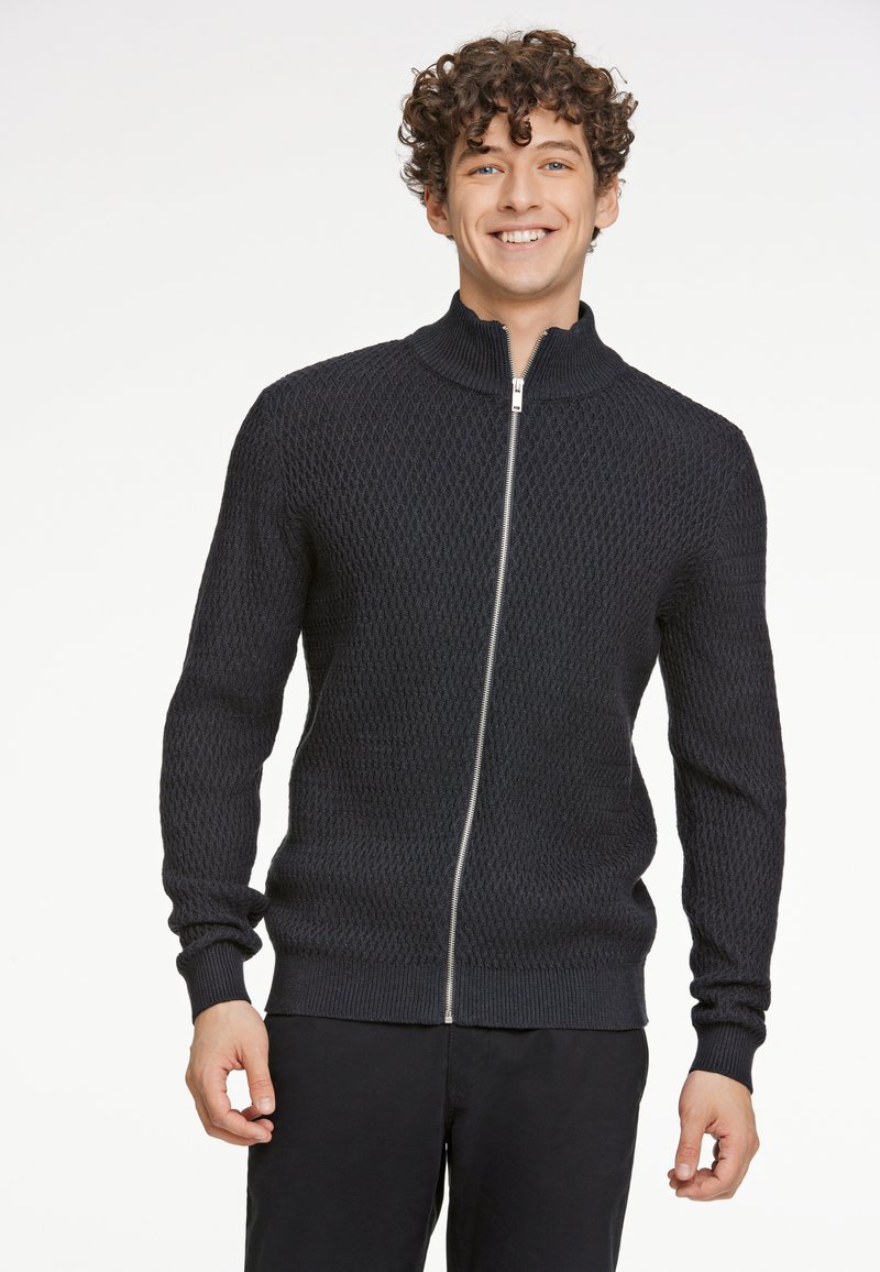 Suéter de punto negro con cremallera y cuello alto, con un patrón texturizado y puños acanalados, combinado con pantalones negros.