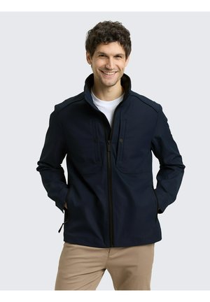 Man glimlachend, draagt een marineblauw jack met rits over een wit shirt, met handen in de zakken en beige broek, staand tegen een lichtgrijze achtergrond.