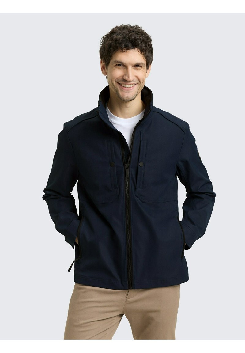 Mann lächelt, trägt eine marineblaue Jacke mit Reißverschluss über einem weißen Hemd, die Hände in den Taschen und beige Hosen, steht vor einem hellgrauen Hintergrund.