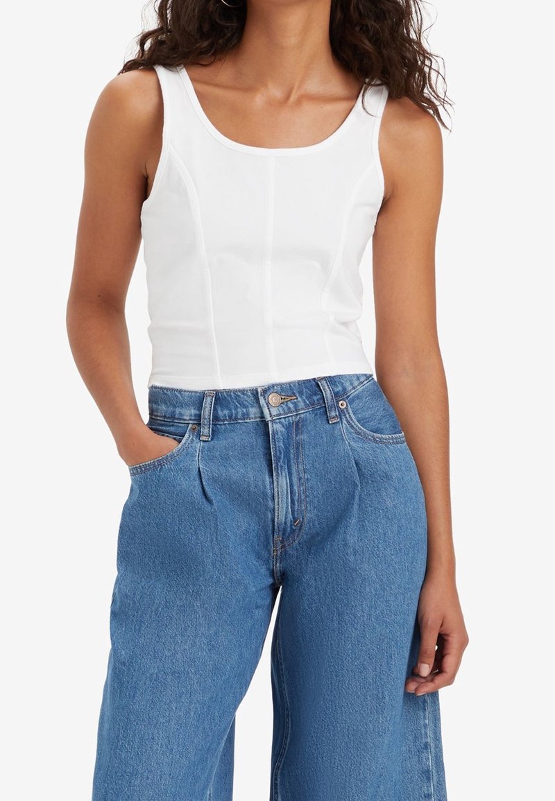 Femme portant un débardeur blanc moulant sans manches et un jean bleu délavé ample taille haute, une main dans la poche.