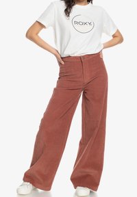 T-shirt léger en coton blanc avec logo circulaire noir ; pantalon en velours côtelé rouge rouille à jambes larges avec une texture côtelée subtile et une couture frontale.