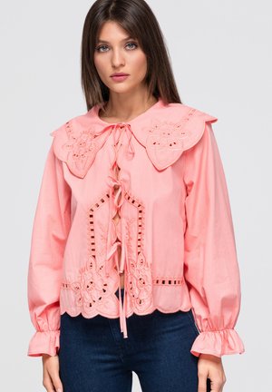 Jeune femme portant un chemisier rose avec une broderie florale, un grand col festonné et un laçage à l'avant, associé à un jean bleu foncé.