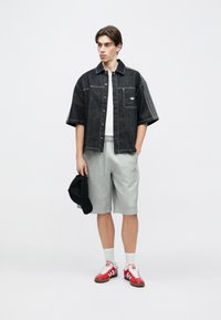 Νεαρός άνδρας με μαύρο τζιν μπουφάν με κοντά μανίκια, λευκό πουκάμισο, γκρι σορτς Adidas, κόκκινα αθλητικά παπούτσια, κρατώντας ένα μαύρο καπέλο σε ένα χέρι.