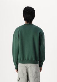 Green sweatshirt with a relaxed fit, round neckline, and dropped shoulders. Cotton blend material. Worn with light grey cargo pants.